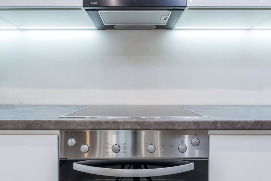 A woman with blonde hair tied in a bun, wearing a white t-shirt and yellow and blue rubber gloves, is using a spray bottle and a blue cloth to clean the interior of a black oven in a modern kitchen. T